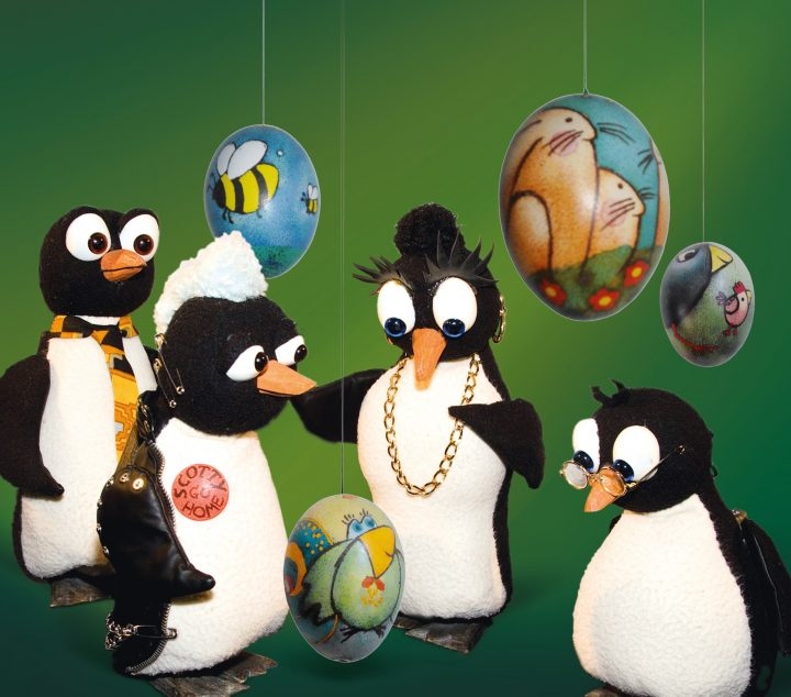 4 Marionetten-Pinguin-Figuren schweben auf moosgrünem Hintergrund. Dazwischen hängen bunt bemalte Ostereier.