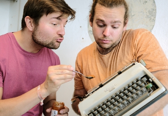 Zwei Trainer für Improvisationstheater füttern eine Schreibmaschine.Die Person links hält den Löffel, die Person trägt die Schreibmaschine wie ein Baby auf dem Arm.