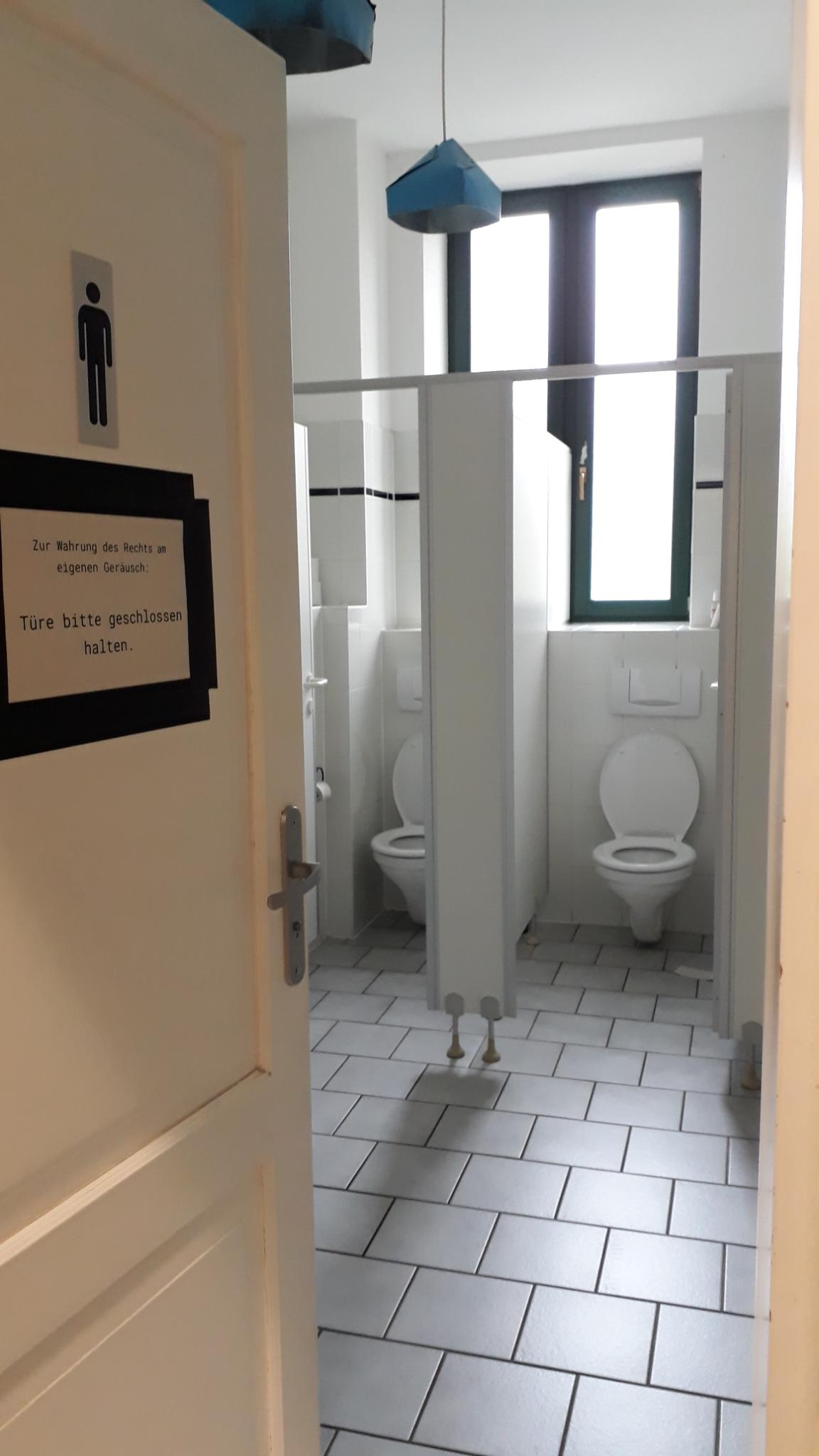 Von außen fotografiert. Eine offene Tür die ins inner des Herren WC-Raums führt. Im Hintergrund zwei offene WC-Kabinen. Blaue Lampenschirme hängen von der Decke.
