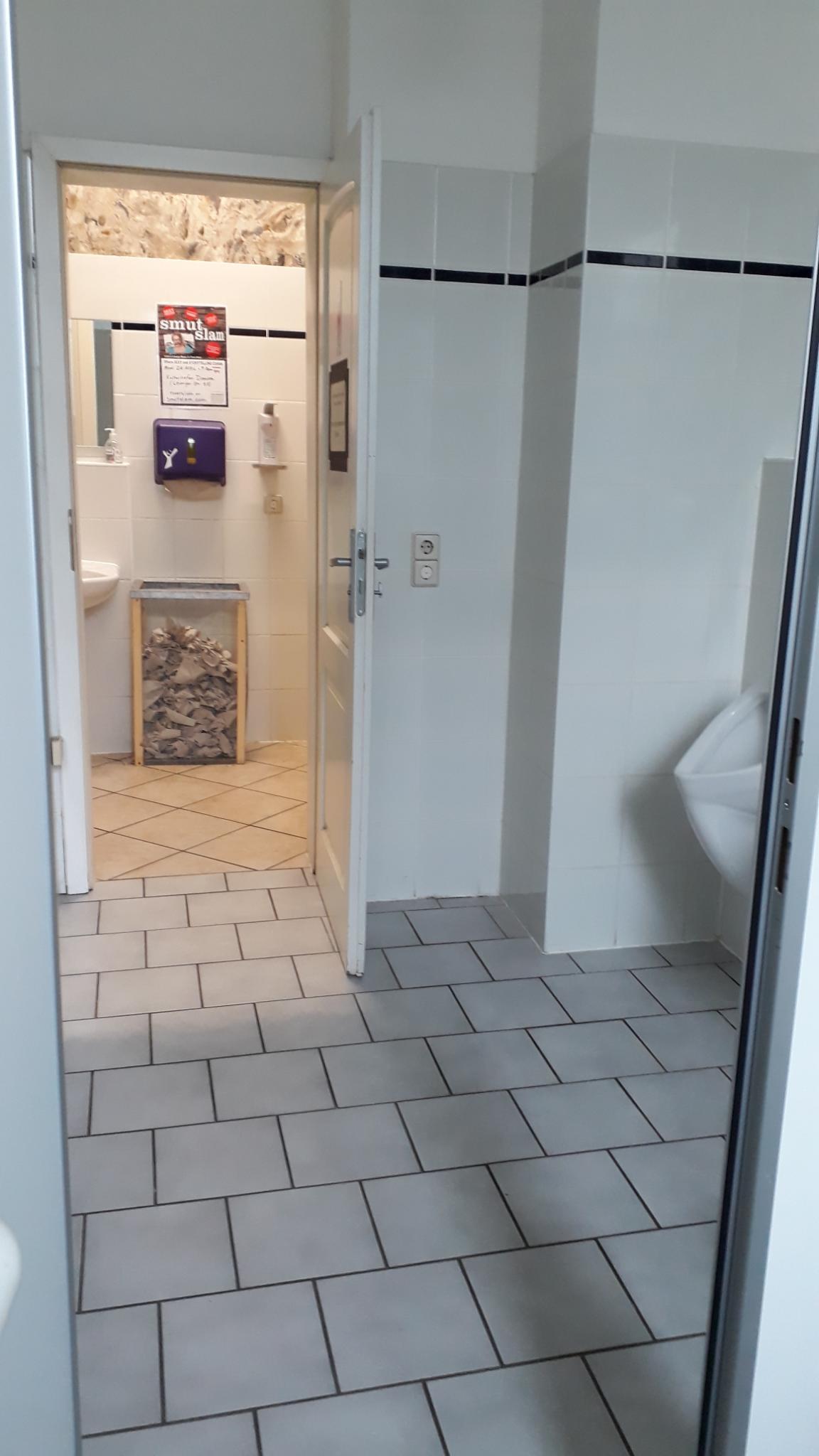 Von innen fotografiert. Links eine offene Tür, welche mit drehknauf von innen verschließbar ist und ins innere des weiß gefließten WC-Raums führt.